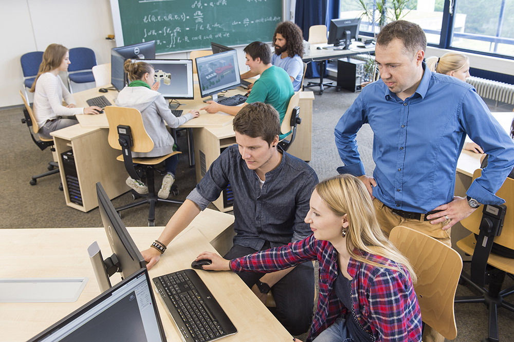 Im Vordergrund arbeiten zwei Studenten am PC zusammen, dabei schaut Ihnen der Prof über die Schulter. Im Hintergrund arbeiten weitere Studenten am PC 