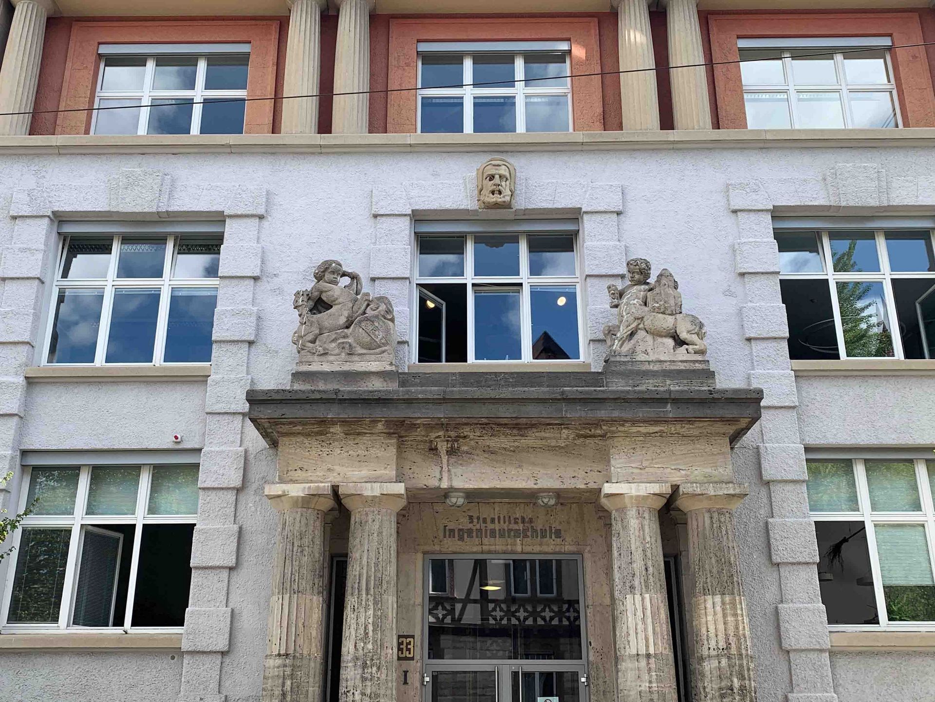Der historische Eingang bei Gebäude 1 am Campus Stadtmitte, über dem Eingang ist der Schriftzug staatliche Ingenieurschule zu lesen, obendrüber sind zwei Steinfiguren. 
