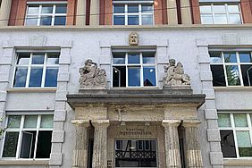 Der historische Eingang bei Gebäude 1 am Campus Stadtmitte, über dem Eingang ist der Schriftzug staatliche Ingenieurschule zu lesen, obendrüber sind zwei Steinfiguren. 