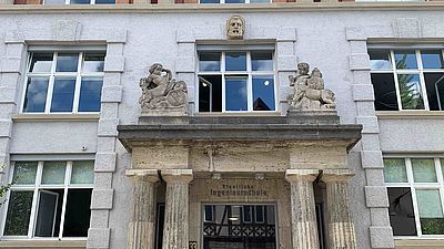 Der historische Eingang bei Gebäude 1 am Campus Stadtmitte, über dem Eingang ist der Schriftzug staatliche Ingenieurschule zu lesen, obendrüber sind zwei Steinfiguren. 