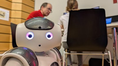 ein Roboter schaut in die Kamera, dahinter sitzen Personen an einem Tisch