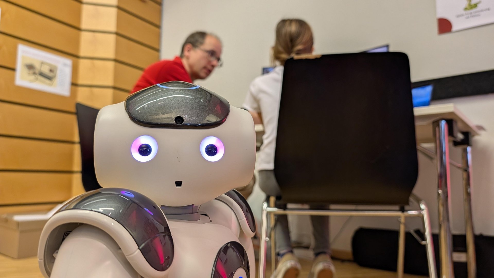 ein Roboter schaut in die Kamera, dahinter sitzen Personen an einem Tisch
