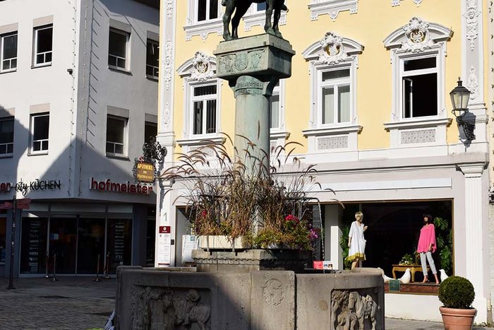 Auf einem weiten Platz steht ein steinerner Brunnen mit Figuren an den Seiten. In der Mitte erhebt sich eine S&auml;ule mit einem Reiter mit Pferd aus Bronze. Der Reiter bl&auml;st in ein Horn.