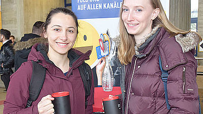 Zwei junge Frauen in weinroten Jacken und Tassen in der Hand.