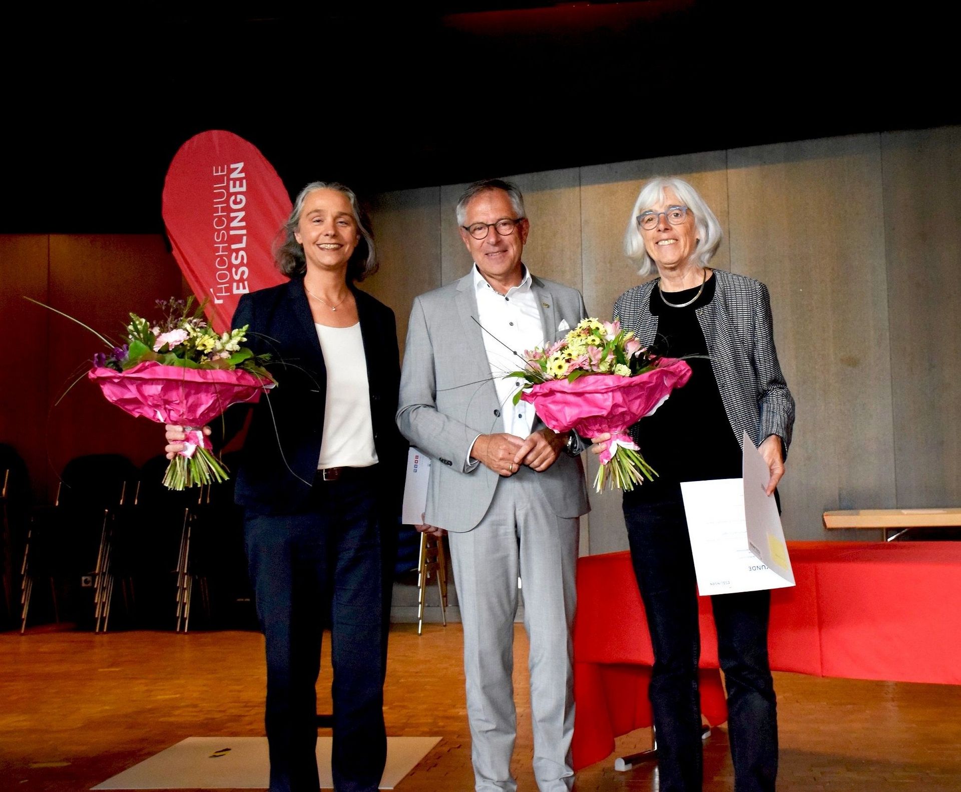 Zwei Frauen mit Blumenstrauß und ein Mann schauen in die Kamera