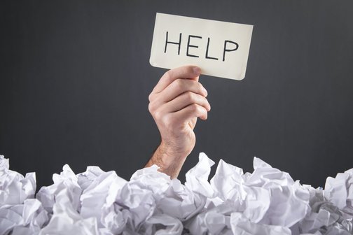 Hand eines Mannes hält ein Schild mit dem Wort "Hilfe" über einen Haufen von zerknäulten Papieren.Male hand holding text Help with a crumpled papers.