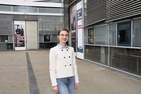 Junge Frau mit Brille und Jacke steht auf dem Campus