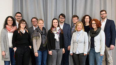 Gruppenfoto von zwölf Menschen vor einem grauen Hintergrund.