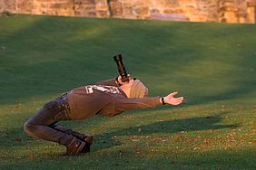Ein Fotograf auf einer Wiese lehnt sich weit in Rückenlage und hält die Kamera in Richtung Himmel.