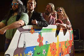 4 Schauspieler in Hippie-Kleidung stehen in Papp-VW-Bus