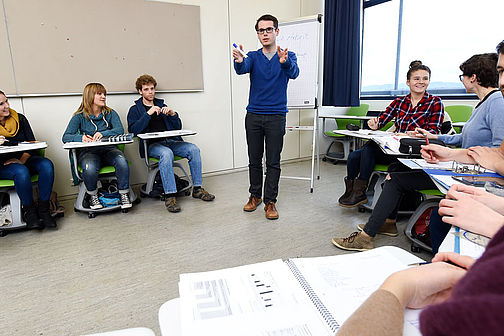 Studierende in den Räumen der Hochschule Esslingen. Einige sitzen auf Stühlen und machen Notizen. Eine Person steht vor einer Flipchart und gestikuliert.