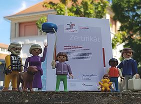 Eine Playmobil-Figuren Familie ist um ein Zertifikat herum angeordnet.