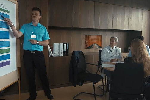 Eine Person steht an der Projektion einer Powerpoint Präsentation, zeigt dabei auf diese. Weitere Personen sitzen im Raum.