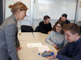 Eine junge Frau und ein junger Mann sitzen an einem Tisch und zeigen einer anderen Frau ein Spielzeug in Fischform.