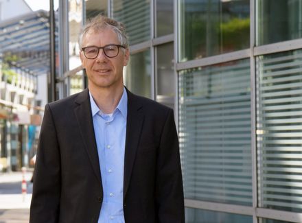 Ein Mann mit Brille, Jacket und blauem Hemd vor einer Glaswand.