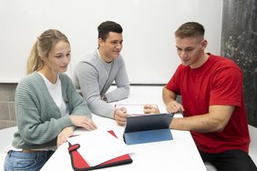 Zwei Männer und eine Frau sitzen an einem Tisch mit Arbeitsmaterial