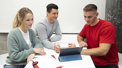 Zwei Männer und eine Frau sitzen an einem Tisch mit Arbeitsmaterial