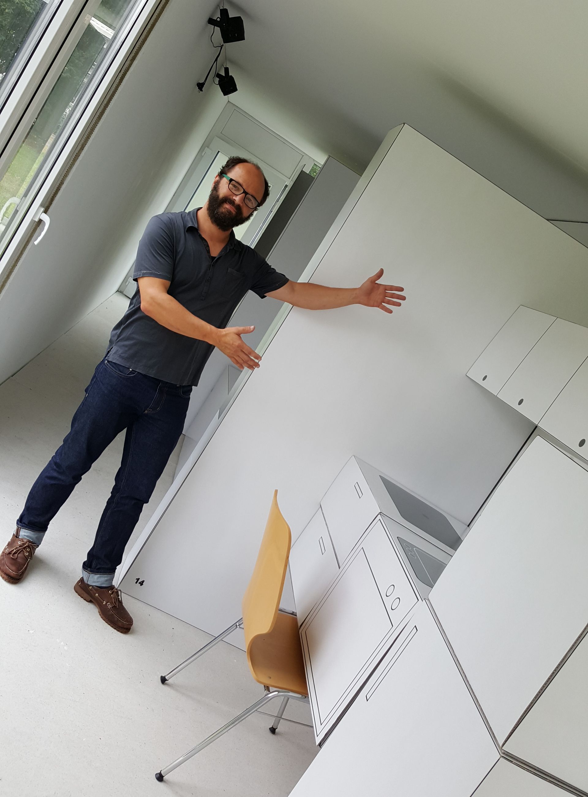 Ein Mann mit Brille steht in einem Mini-Haus und zeigt auf eine Küche aus Pappe.