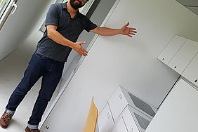 Ein Mann mit Brille steht in einem Mini-Haus und zeigt auf eine Küche aus Pappe.