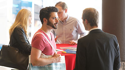 Ein Student steht an einem roten Stehtisch und spricht mit einem Mann. Im Hintergrund stehen eine Frau und ein Mann.