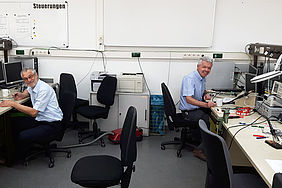 Ein Raum in dem links und rechts jeweils ein Professor vor einem Schreibtisch sitzt. 