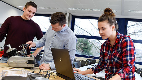 Eine Gruppe Studierende stehen am Tisch und arbeiten am Laptop