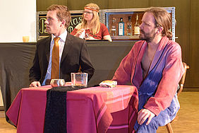 Schauspieler sitzen an einem Tisch an einer Bar, im Hintergrund Barbesitzerin Barbara Ann