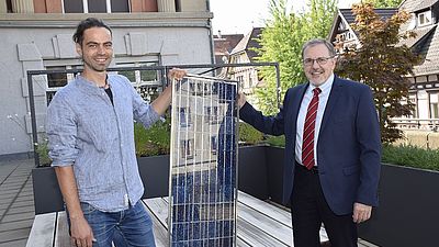 Zwei Männer stehen mit einem Solarpanel in der Hand auf der Terrasse der Hochschule