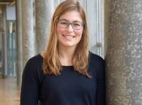 Eine junge Frau mit Brille lehnt an einer Säule.