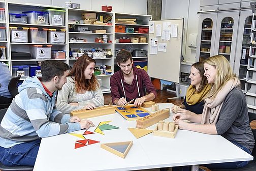 Studierende sitzen um einen Tisch in der Bildungswerkstatt herum. Auf dem Tisch liegen Form und Geometriespiele.
