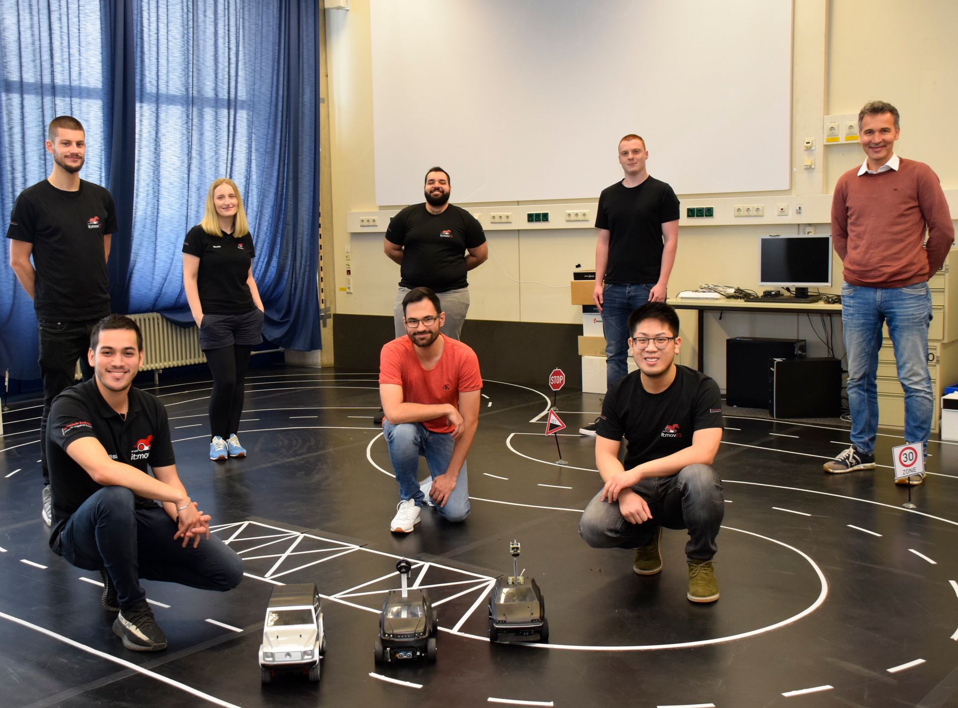 Acht Personen auf einer Strecke für Modellautos. Drei Modellautos stehen vor der Gruppe auf dem Boden. 