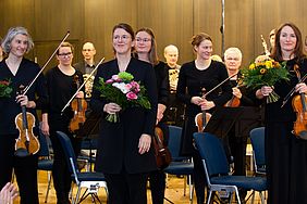 Drei Frauen mit Blumensträußen vor einem Orchester