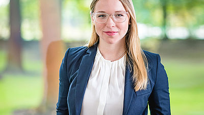 Eine junge Frau mit langen blonden Haaren, brille und blauem Blazer in einer grünen Landschaft. 