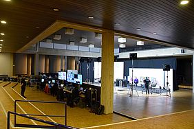 Überblicksbild über das Fernsehstudio in der Aula. Im Vordergrund mit Mischpulten und Monitoren, im Hintergrund mit Lichtern, Kameras und Bühne.