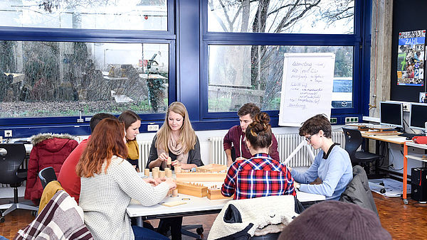 7 Studenten sitzen am Tisch im Computerraum und sind an einer brettspielähnlichen Gruppenarbeit.