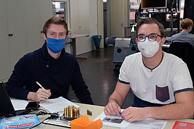 Zwei junge Männer mit Mundschutz sitzen an einem Tisch.