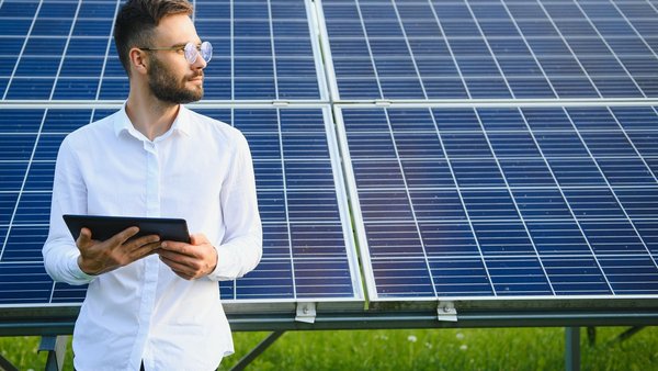 Bearded male engineer using tablet to control photovoltaic panels remotely during work on solar farm on sunny day.