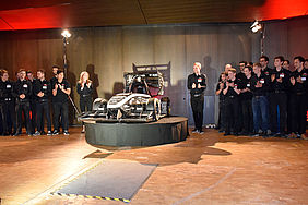 Auf einer Bühne steht in der Mitte ein Rennauto. Links und rechts daneben stehen mehrere Menschen.