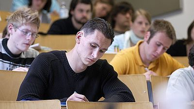 Studierende in der Vorlesung- Fokus auf einen schreibenden Studierenden 
