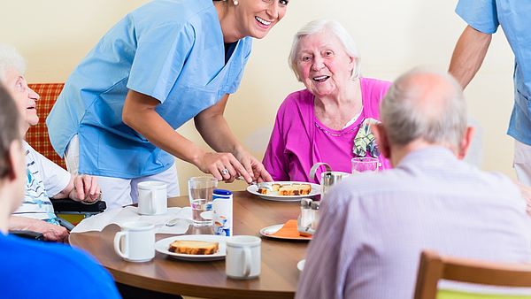 Eine Gruppe von älteren Personen die zusammen am Frühstücktisch sitzen. Eine Krankenhelferin schneidet das Essen Mundgerecht 