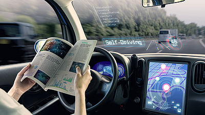 Eine Person sitzt in einem autonom fahrenden Auto und liest in einem Heft.