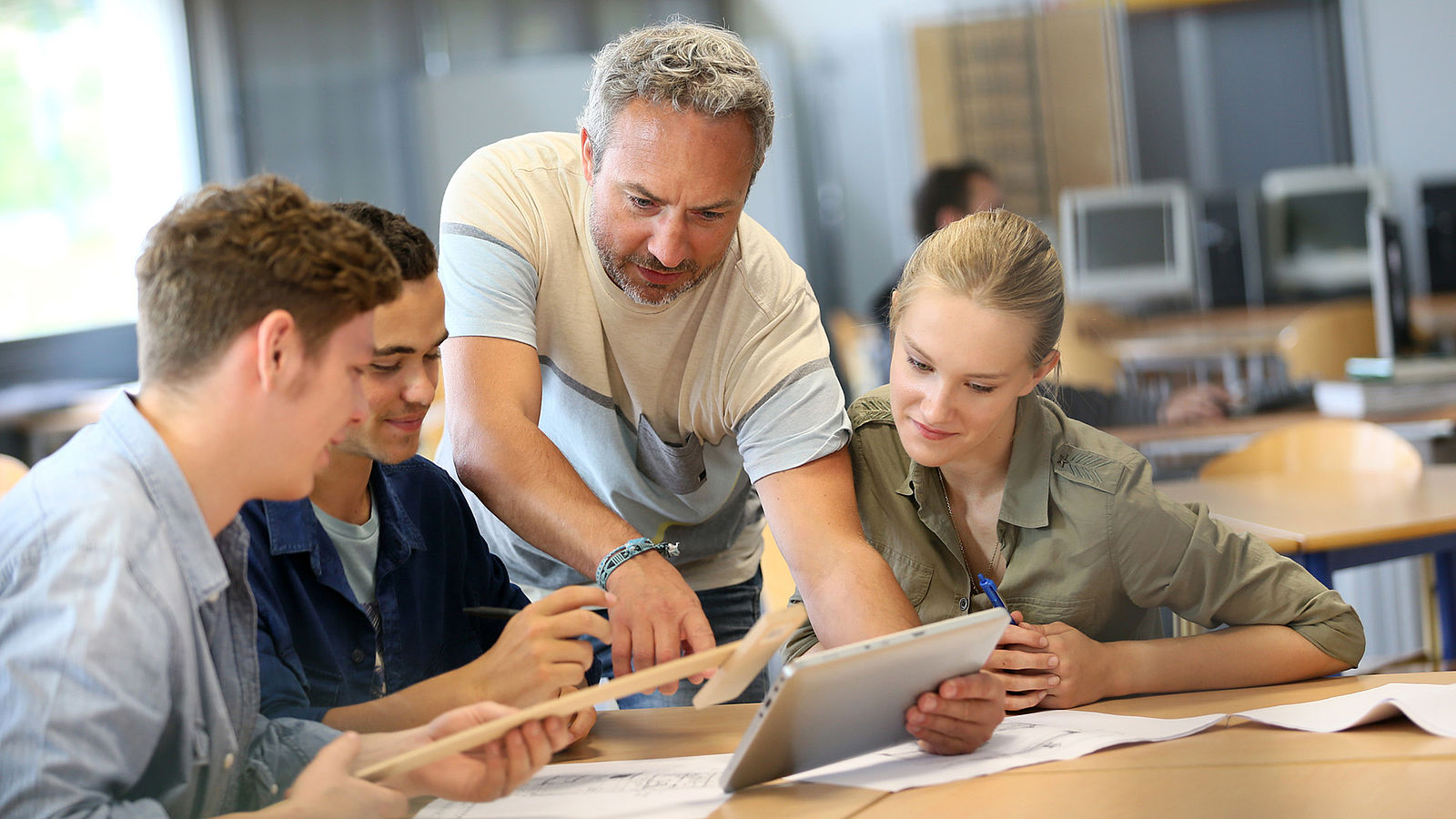 An NWT teacher stands at a desk with his students and looks at a tablet.