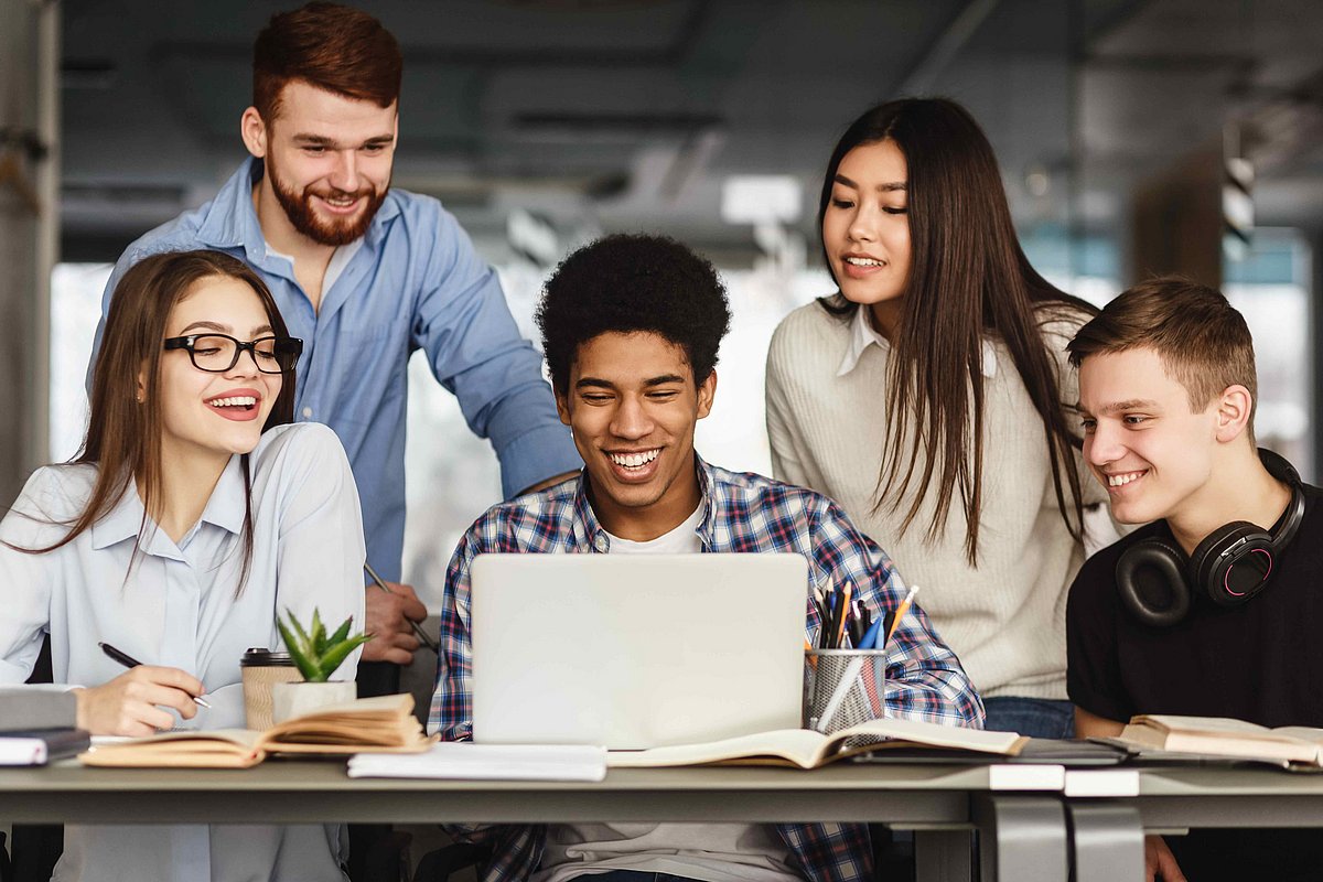 Fünf Studierende sitzen zusammen an einem Laptop und lachen.