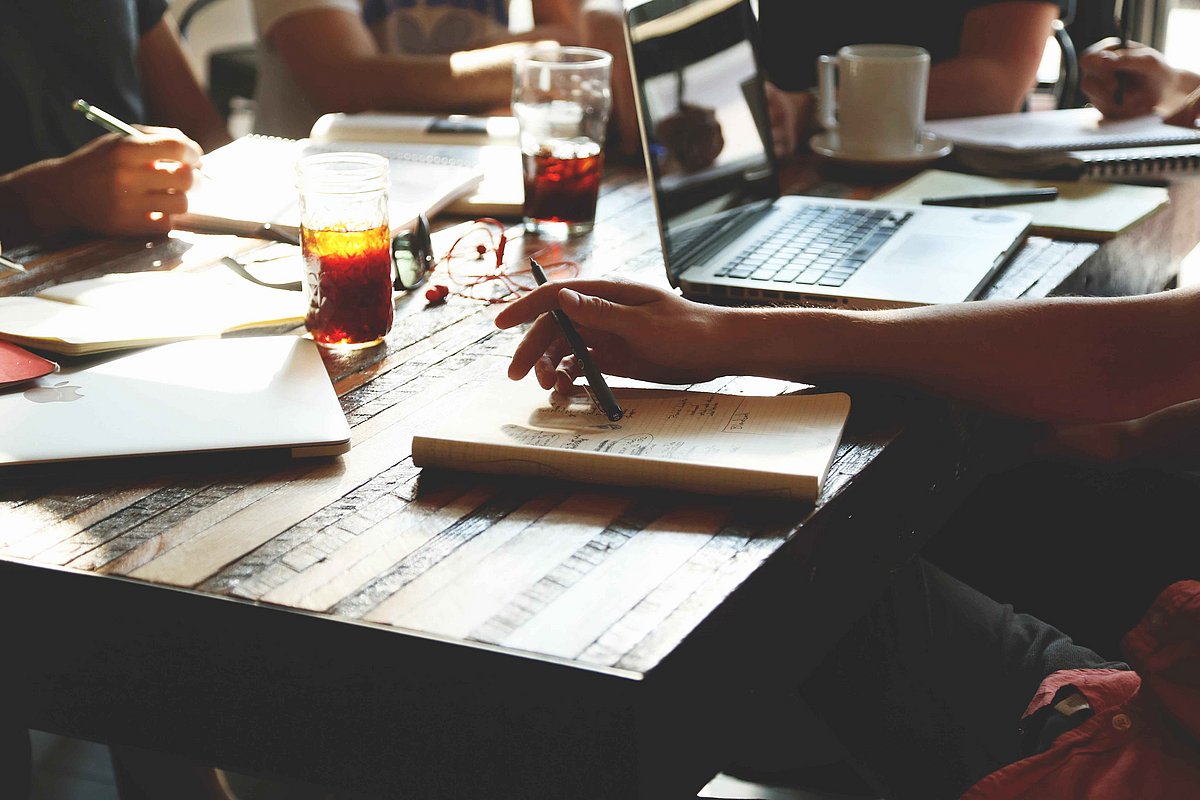 Auf einem Tisch liegen Dokumente und ein Laptop, zwei Personen sitzen am Tisch und arbeiten.