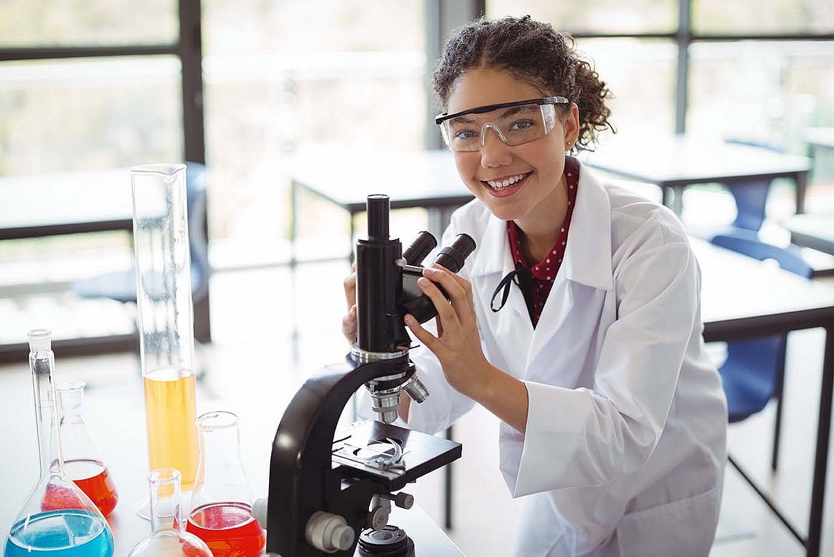 Ein junges Mädchen in einem Laborkittel steht an einem Mikroskop und lächelt.