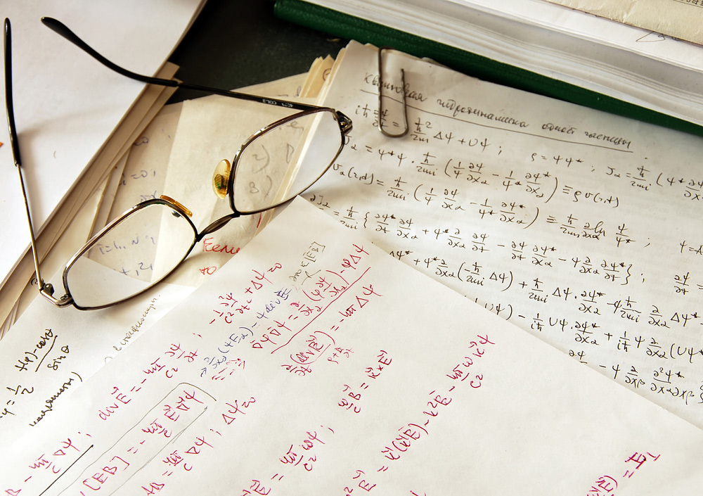 Auf einem Tisch liegen mehrere Blätter mit physikialischen Formeln, darauf liegt eine Brille.