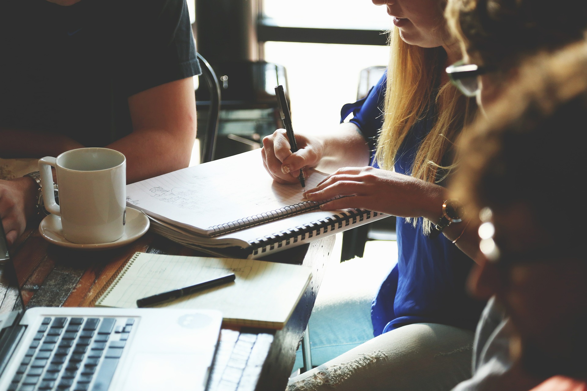 Studierende sitzen gemeinsam am Tisch. Auf dem Tischen liegen Arbeitsmaterialien wie Laptop und Collegeblock. Eine Studentin schreibt. 