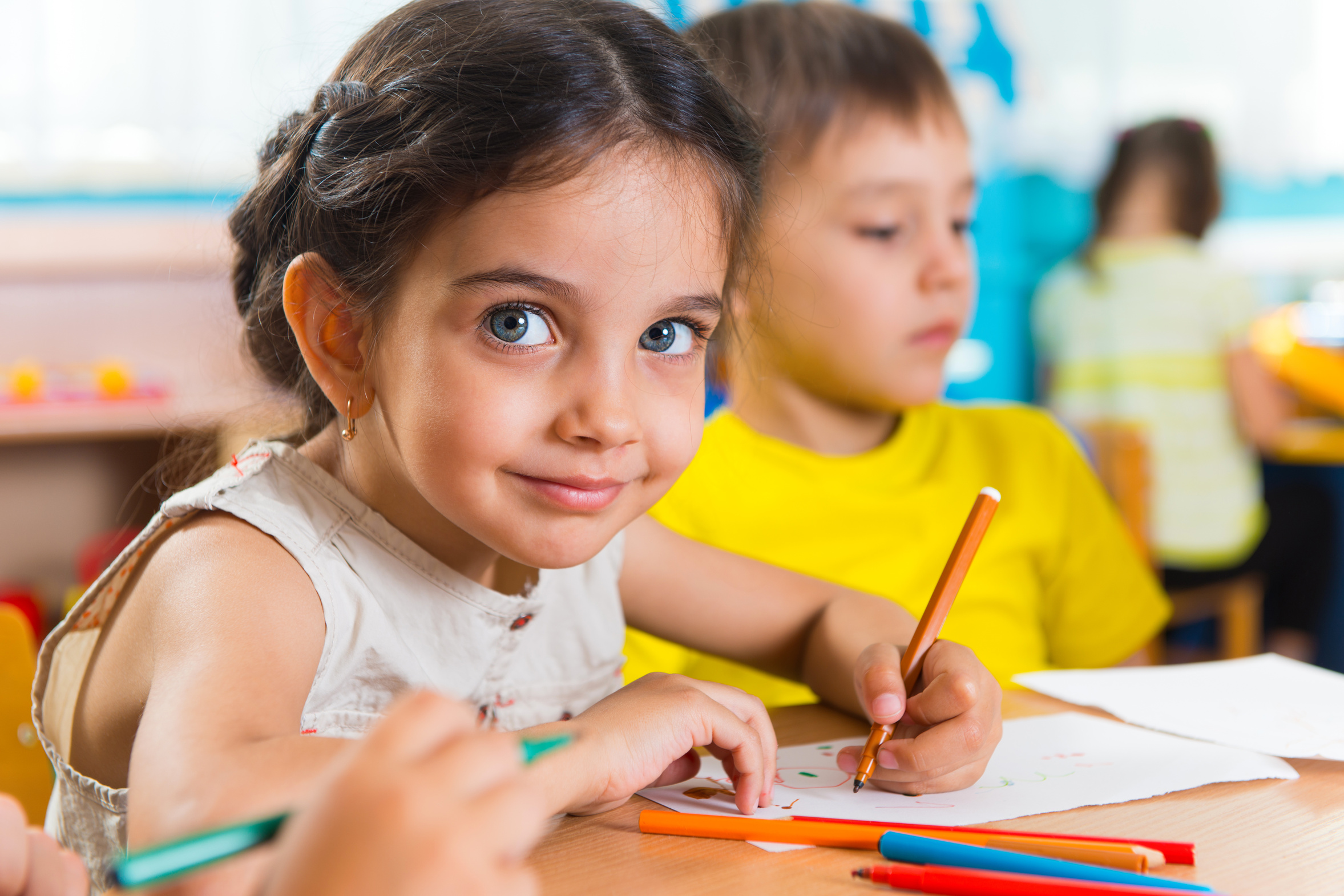 Foto von zwei Kindern am Tisch sitzend. Ein Kind schaut in die Kamera während es auf einem Blatt Papier malt.