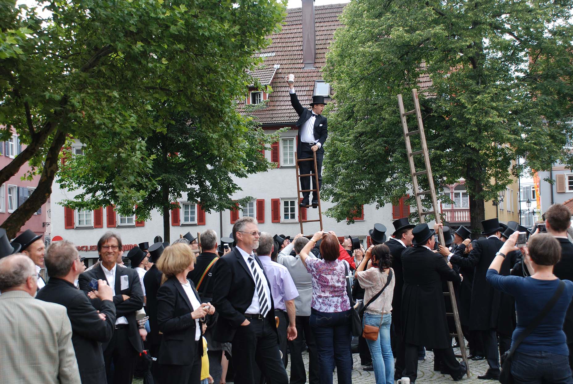 Über einer Menschenmenge steht ein Student auf einer Leiter und hebt seinen Bierkrug.
