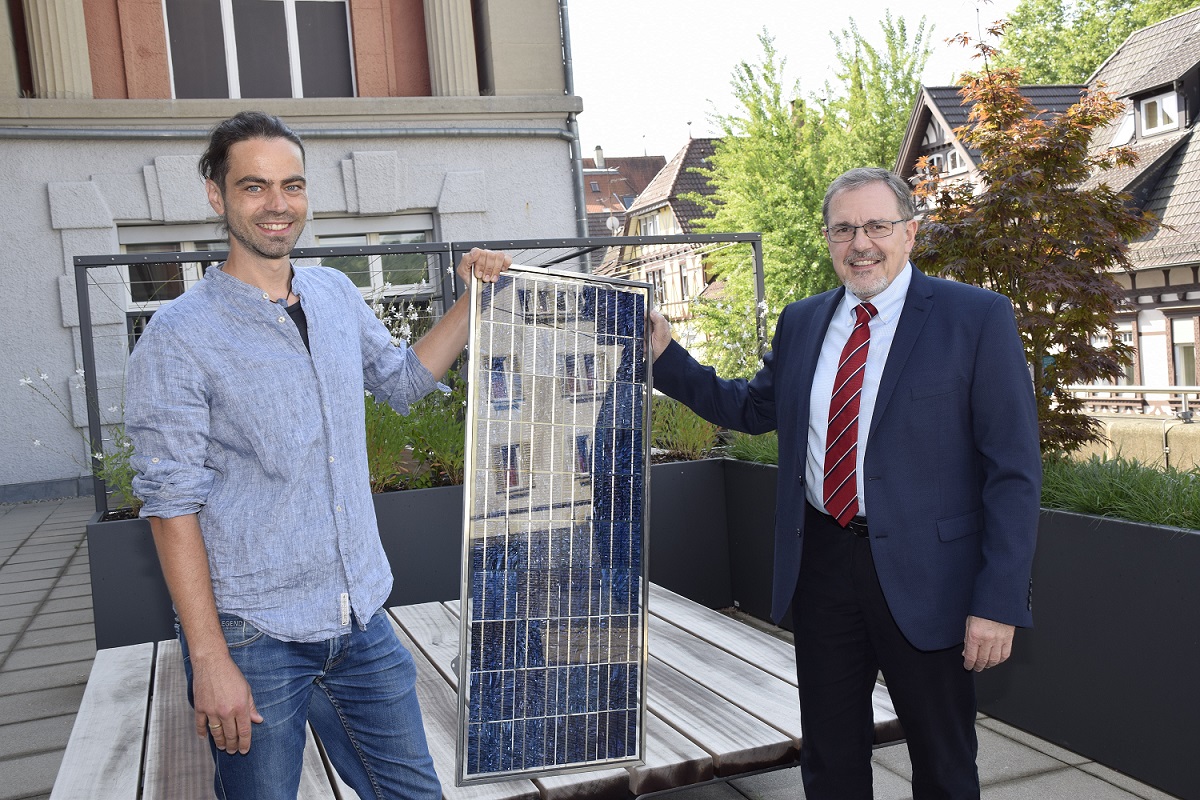Zwei Männer stehen mit einem Solarpanel in der Hand auf der Terrasse der Hochschule